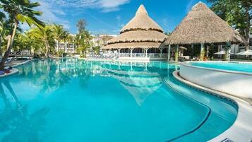 A swimming pool with two thatched-roof structures, palm trees, and lounge chairs.