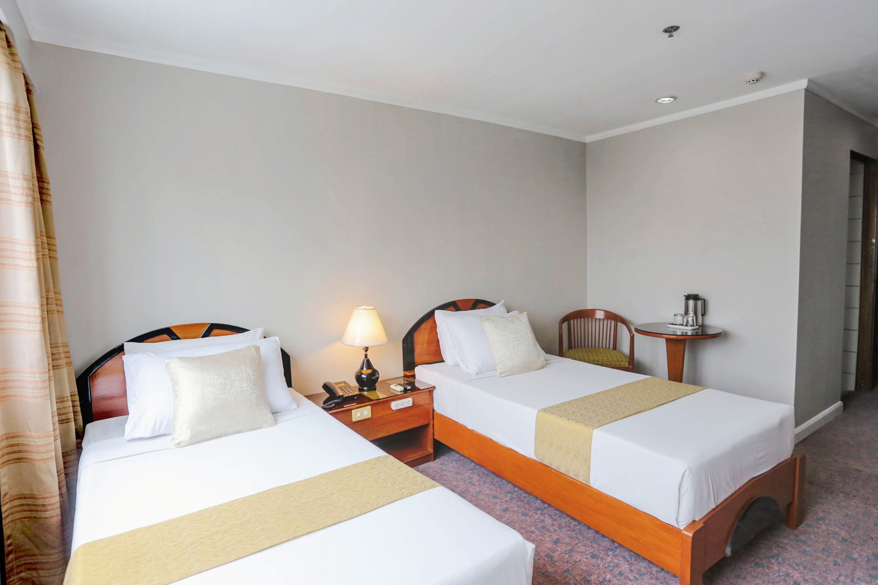 A hotel room with two beds, a nightstand with a lamp, a small table with a coffee maker, and a door leading to another room.