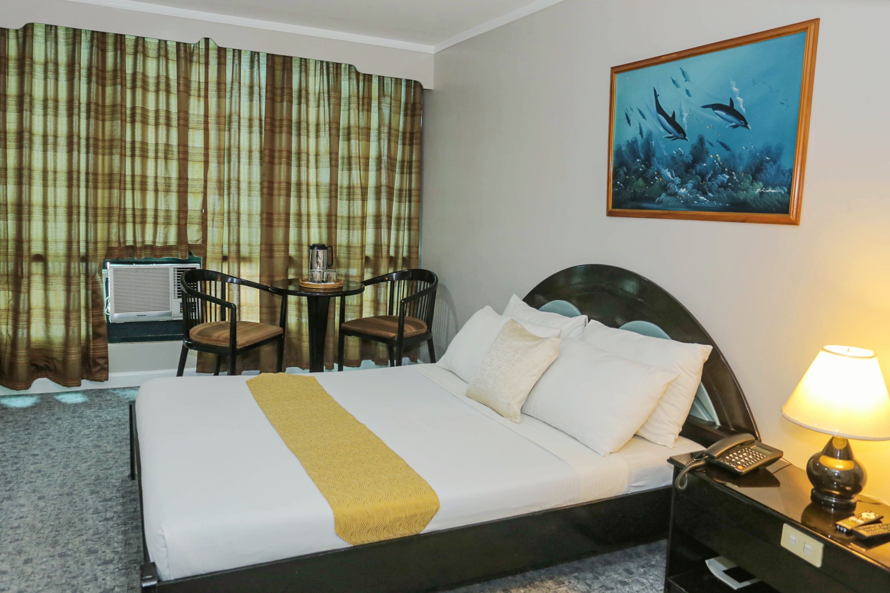 A hotel room with a bed, a desk with two chairs, a painting of dolphins, and a nightstand with a lamp.