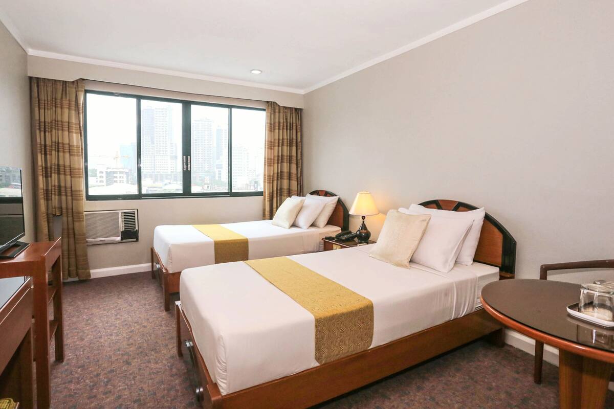 A hotel room with two beds, a desk, a chair, a table, and a large window with a city view.