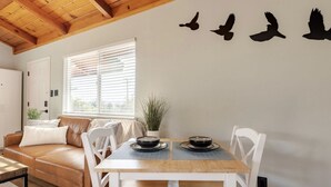 Un comedor con mesa de madera y sillas blancas, una ventana con persianas, un sofá de cuero marrón y un adhesivo mural con aves.