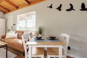 Un comedor con mesa de madera y sillas blancas, una ventana con persianas, un sofá de cuero marrón y un adhesivo mural con aves.