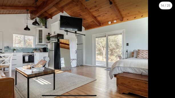 Una habitación amplia con una cama grande, techo de madera y una zona de cocina con armarios blancos y un refrigerador negro.