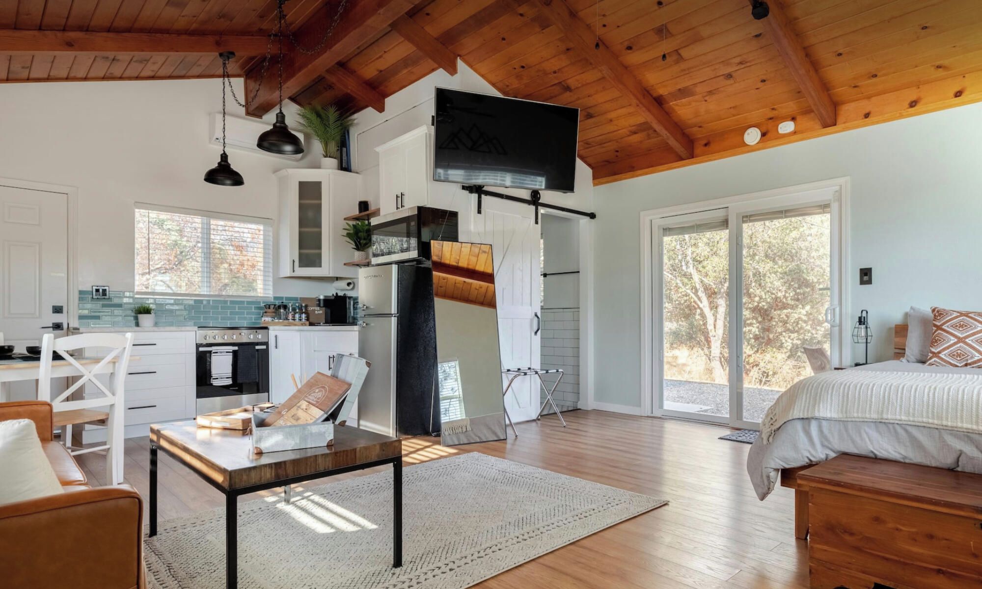 A spacious room with a large bed, a wooden ceiling, and a kitchen area with white cabinets and a black refrigerator.