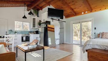 A spacious room with a large bed, a wooden ceiling, and a kitchen area with white cabinets and a black refrigerator.