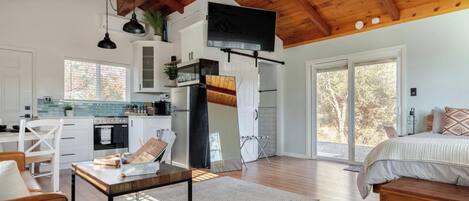 A spacious room with a large bed, a wooden ceiling, and a kitchen area with white cabinets and a black refrigerator.