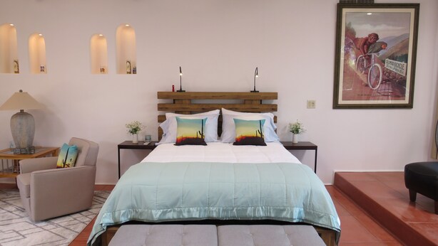 Une chambre à coucher avec un grand lit, une tête de lit en bois, des tables de chevet avec des lampes, une chaise, un banc et un tableau encadré au mur.
