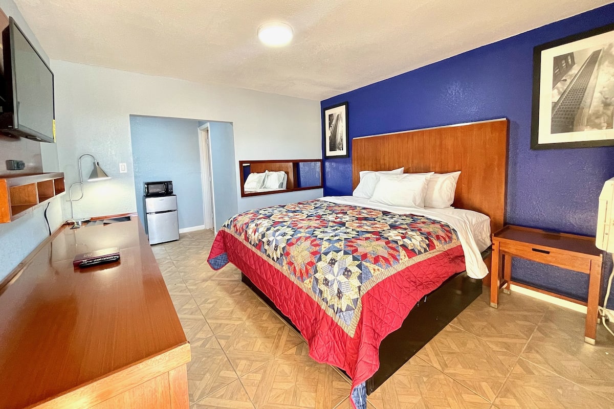A hotel room with a bed, a desk, a chair, a TV, a microwave, and a refrigerator.