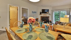 A dining area with a wooden table, green placemats, and a vase of flowers. There is a television above the fireplace and a sofa in the background. - The Woods Resort and Spa Killington: 2 BR / 2 BA Renovated Condos w/ Great Amenities V7 (Killington)