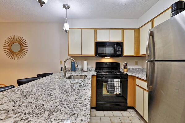 A kitchen with granite countertops, a black stove, a stainless steel refrigerator, a microwave, and wooden cabinets.