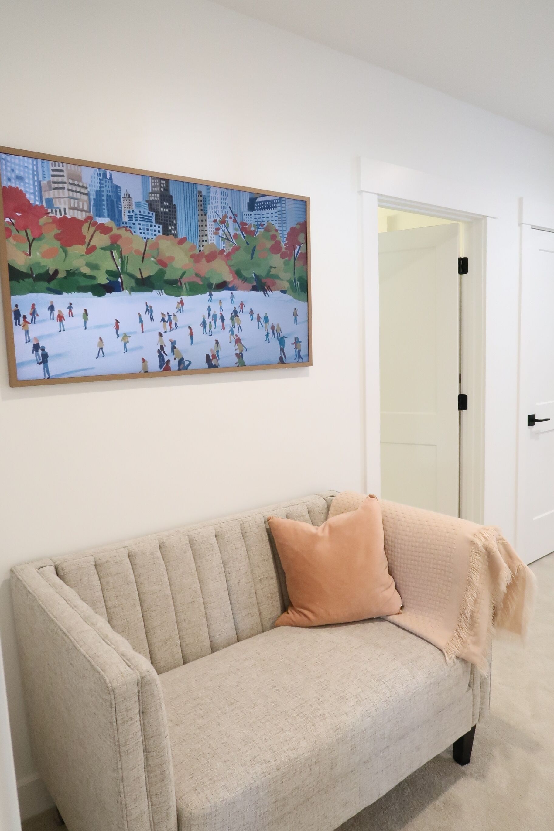 A beige sofa with a single cushion, a framed painting of a cityscape with people, and a white door in the background.