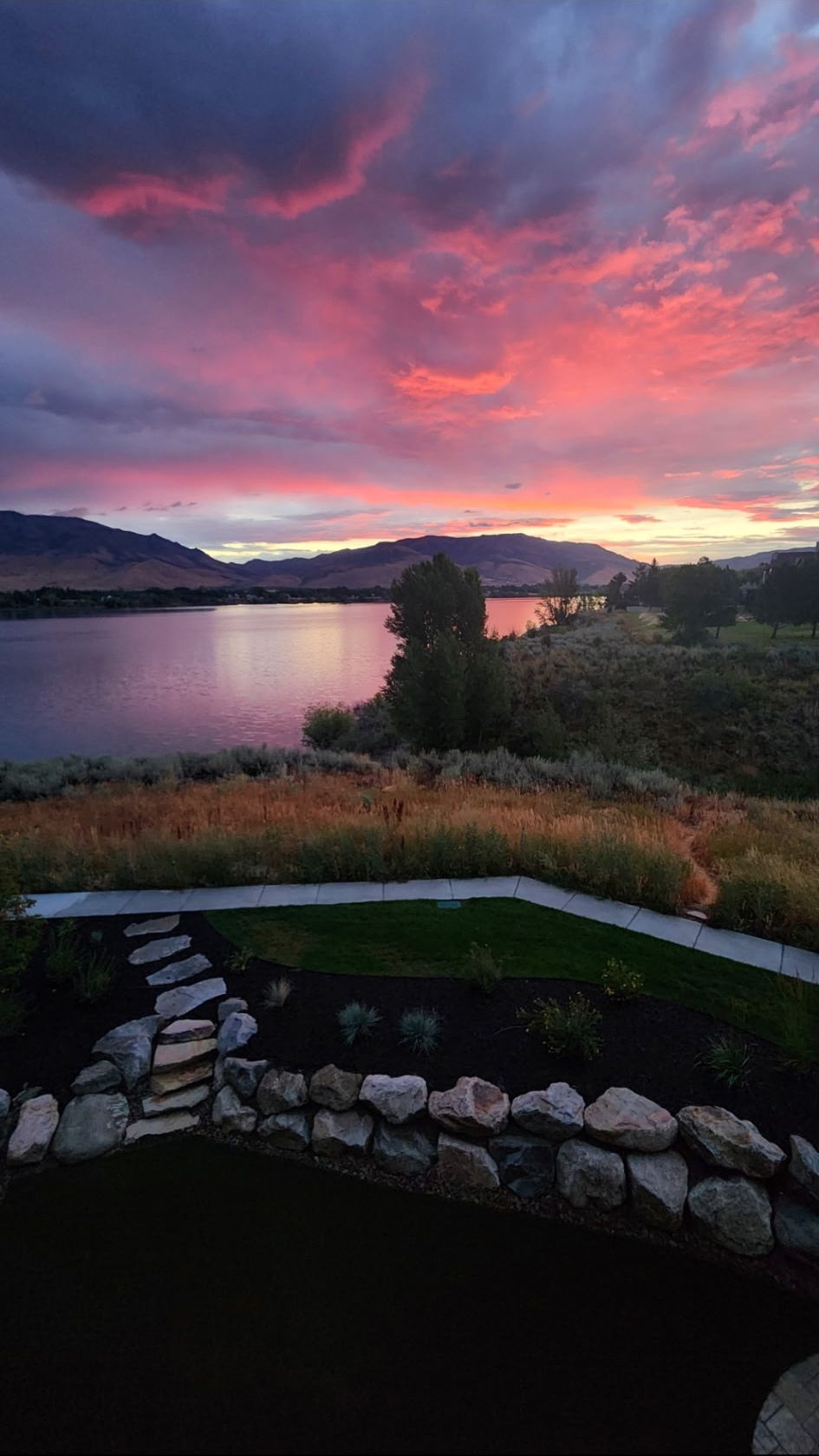 Un paysage au bord d’un lac, avec un sentier de pierres menant à une zone herbeuse, le tout entouré de collines et surplombé par un ciel au coucher du soleil.