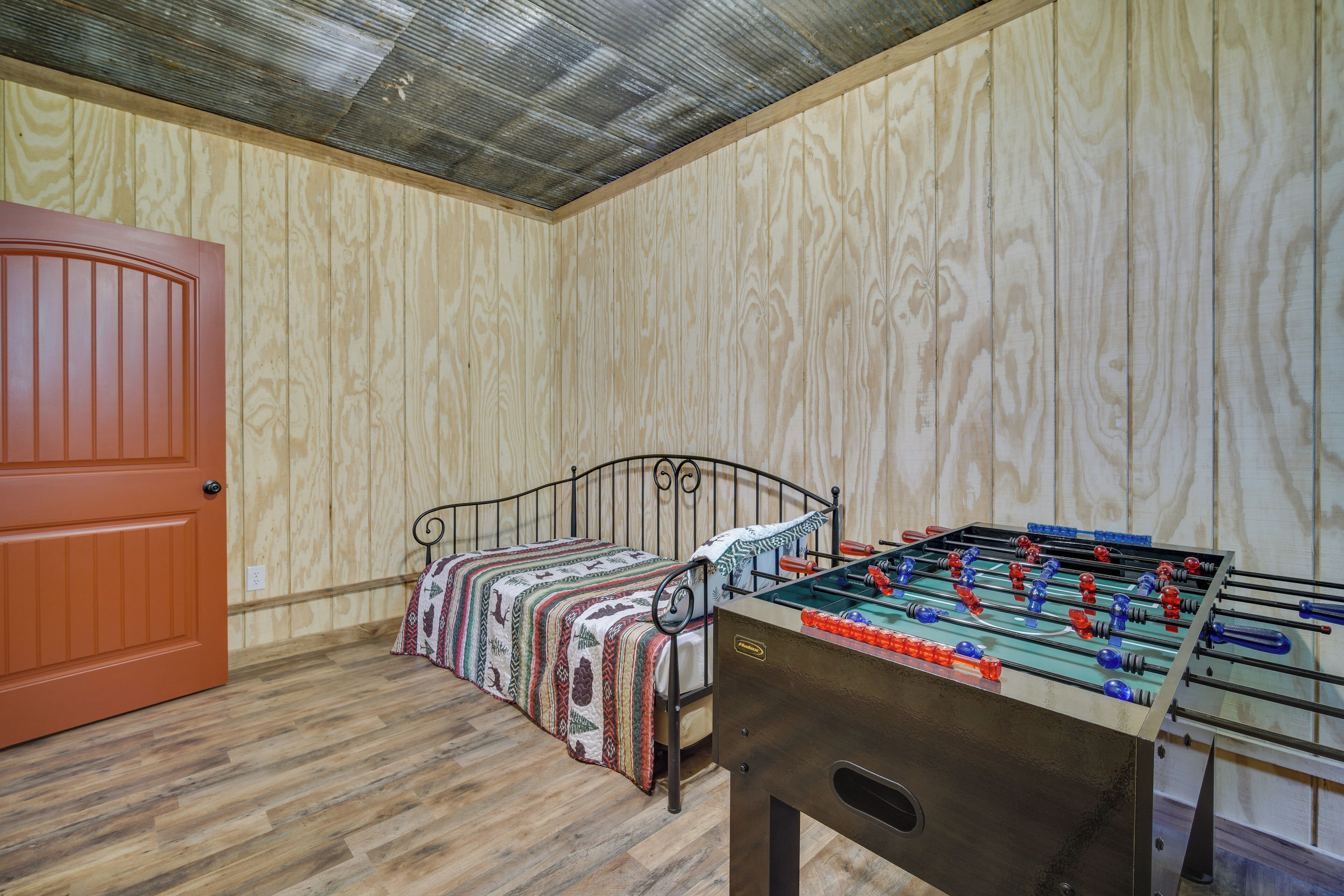 A room with a wooden floor, a bed with a patterned blanket, a red door, and a foosball table.