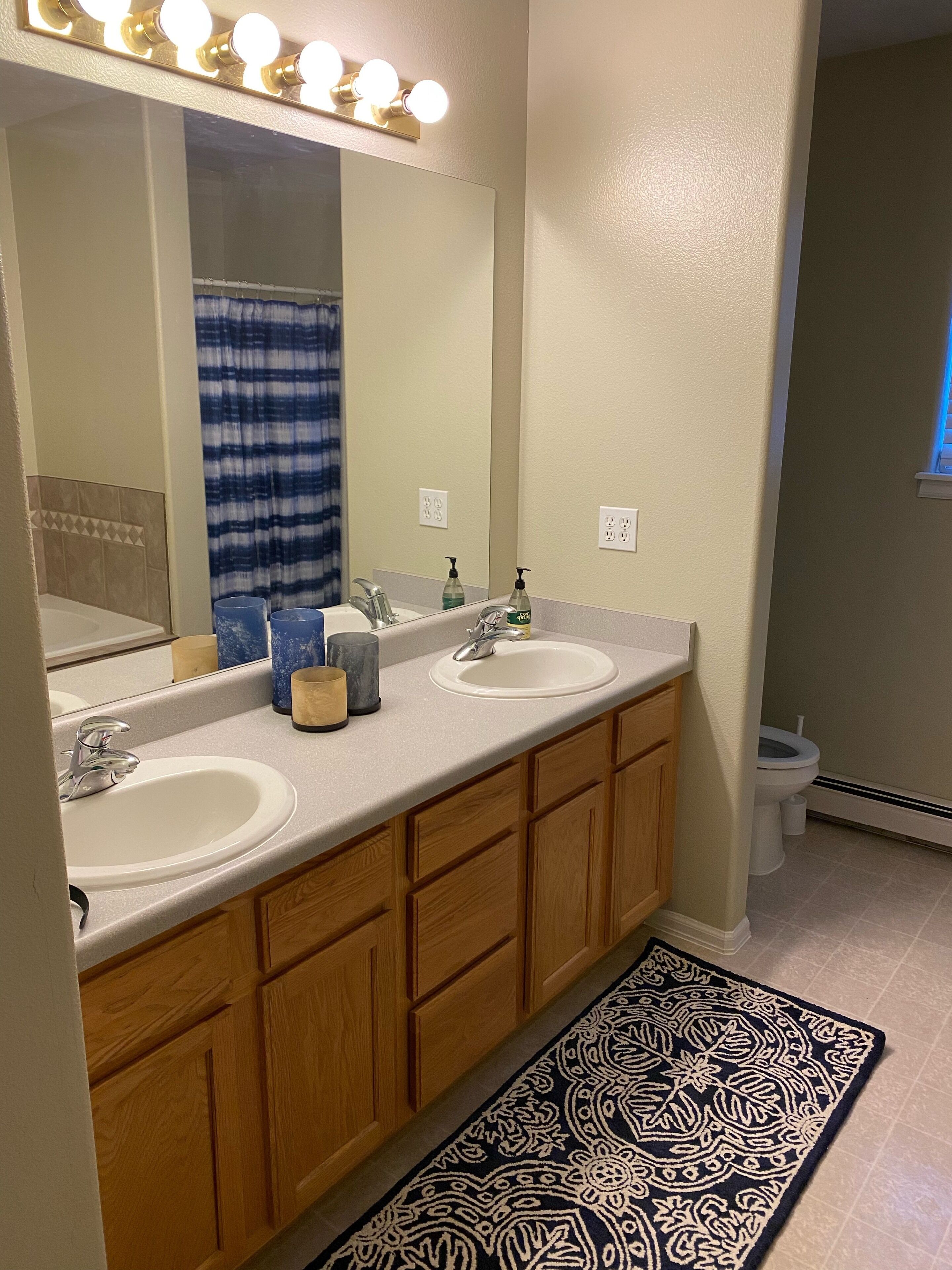 Une salle de bain avec un meuble-lavabo à deux vasques, un grand miroir avec éclairage intégré, un rideau de douche bleu et un tapis à motifs.