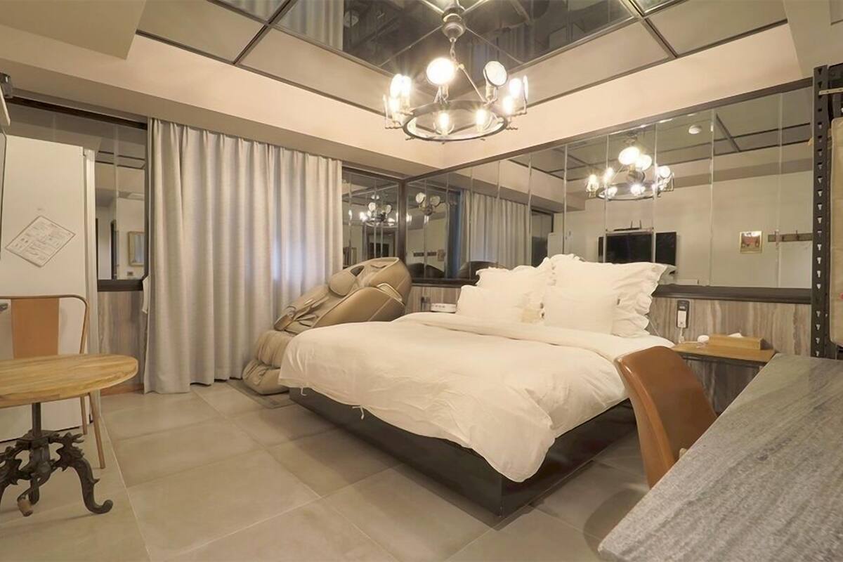 A modern bedroom with a large bed, a desk, and a chair. There is a mirror on the wall and a ceiling light fixture.