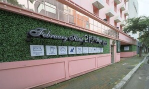 The exterior of the February Hotel 2nd Stay, featuring a green wall with various icons indicating amenities such as a bed, parking, and Wi-Fi.