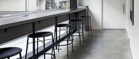 A modern bar area with a long counter and four stools, a large window with a view of buildings, and a concrete floor.