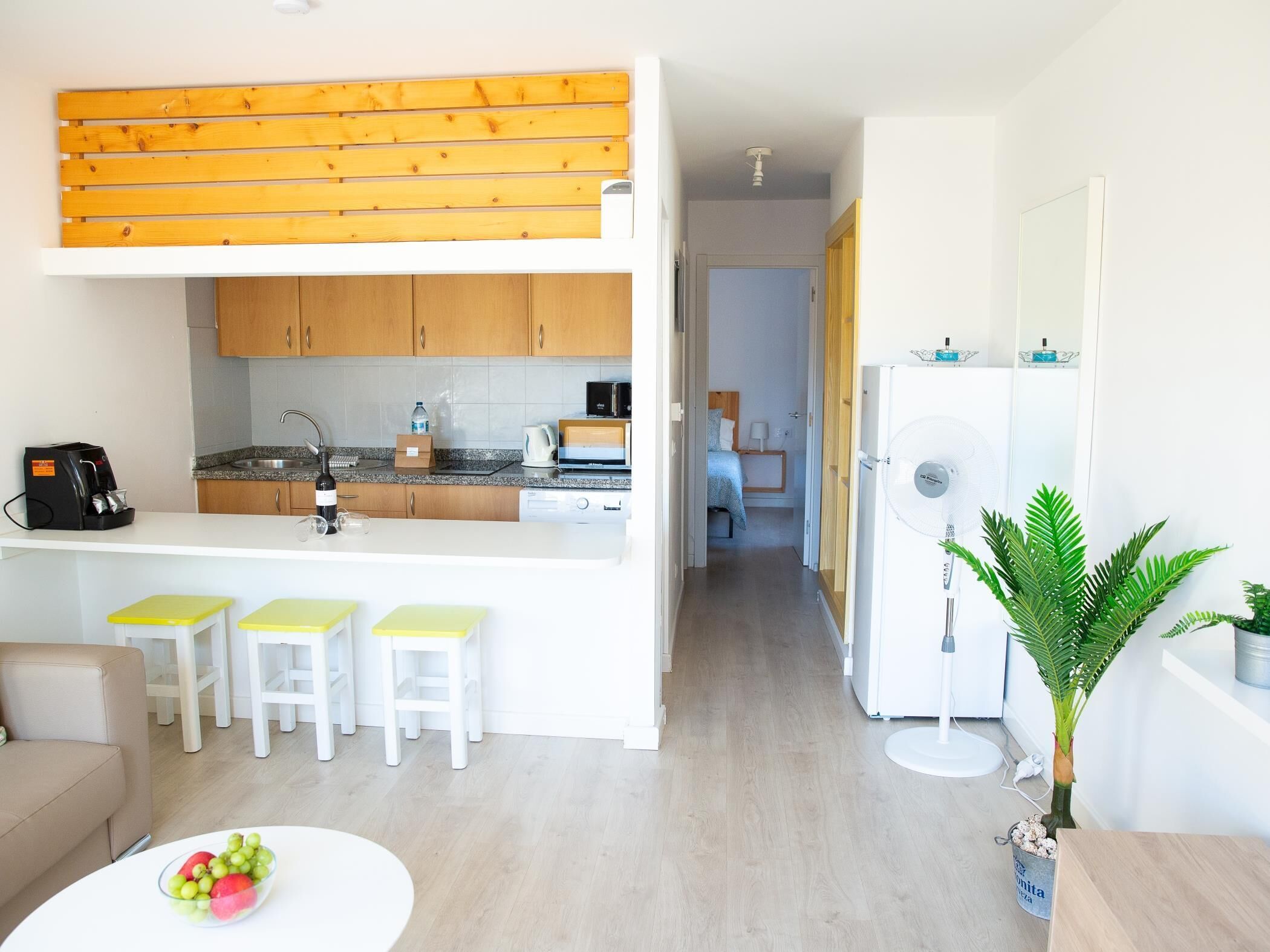 A compact kitchen with wooden cabinets, a coffee machine, and a refrigerator. There are three yellow stools at the counter, and a dining table with fruit. A plant is in a bucket on the floor.