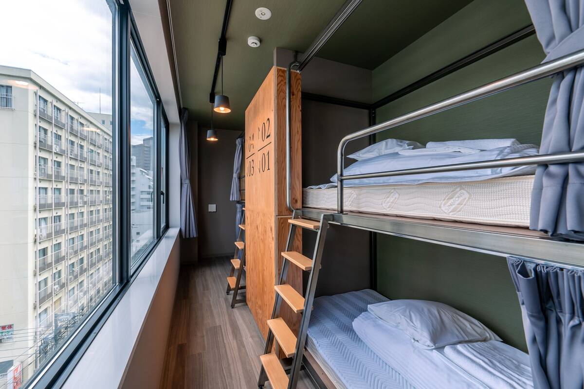 A compact hotel room with bunk beds, a wooden ladder, and a view of a cityscape through large windows.