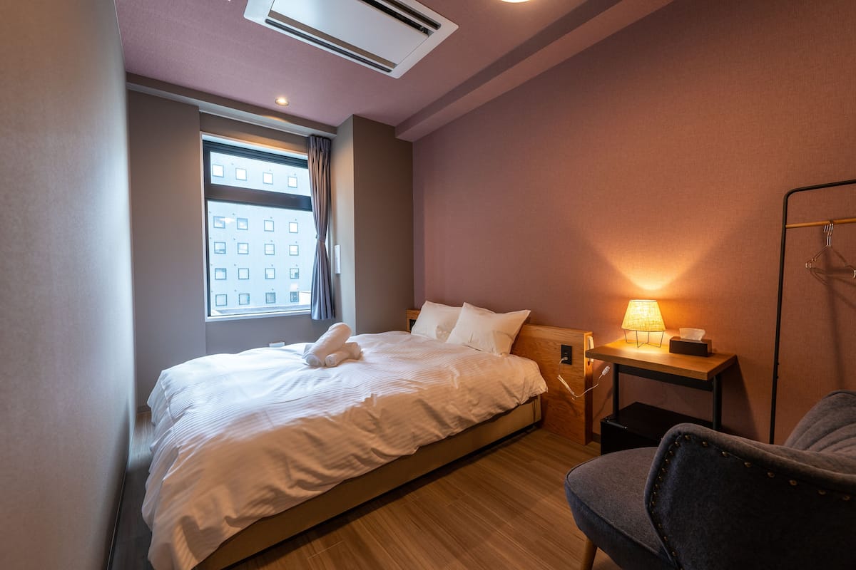 A hotel room with a bed, a chair, a nightstand with a lamp, and a window with curtains.