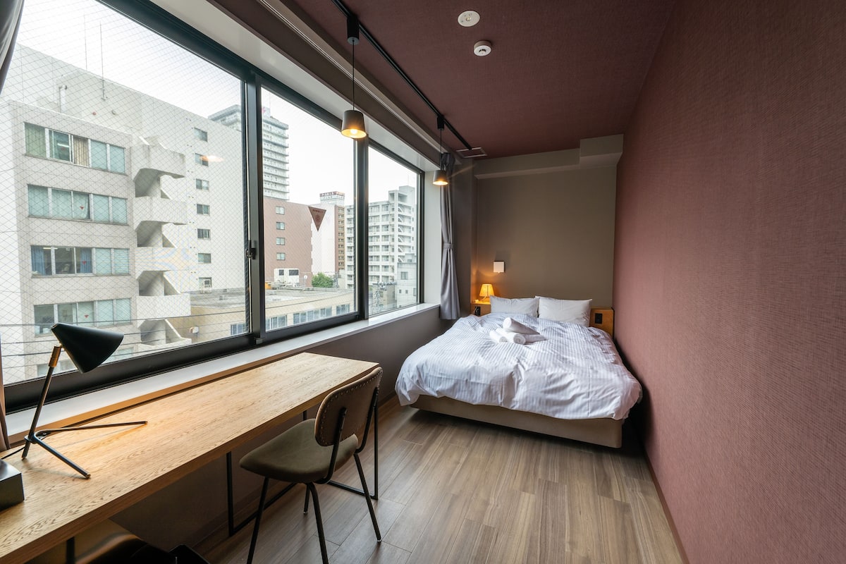 A modern hotel room with a large window, a wooden desk, a bed, and a chair.