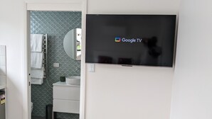 A room with a mounted flat-screen TV displaying the Google TV logo, a white door leading to a bathroom with a tiled backsplash, and a wooden floor. - Secluded Pool House getaway- 500m to patrolled Shelly Beach (Shelly Beach)