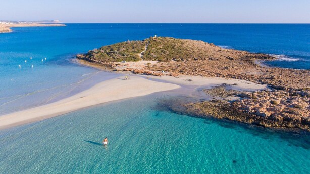 Luchtbeeld van een strand met helder turquoise water, een zandstrand en rotsachtig terrein.