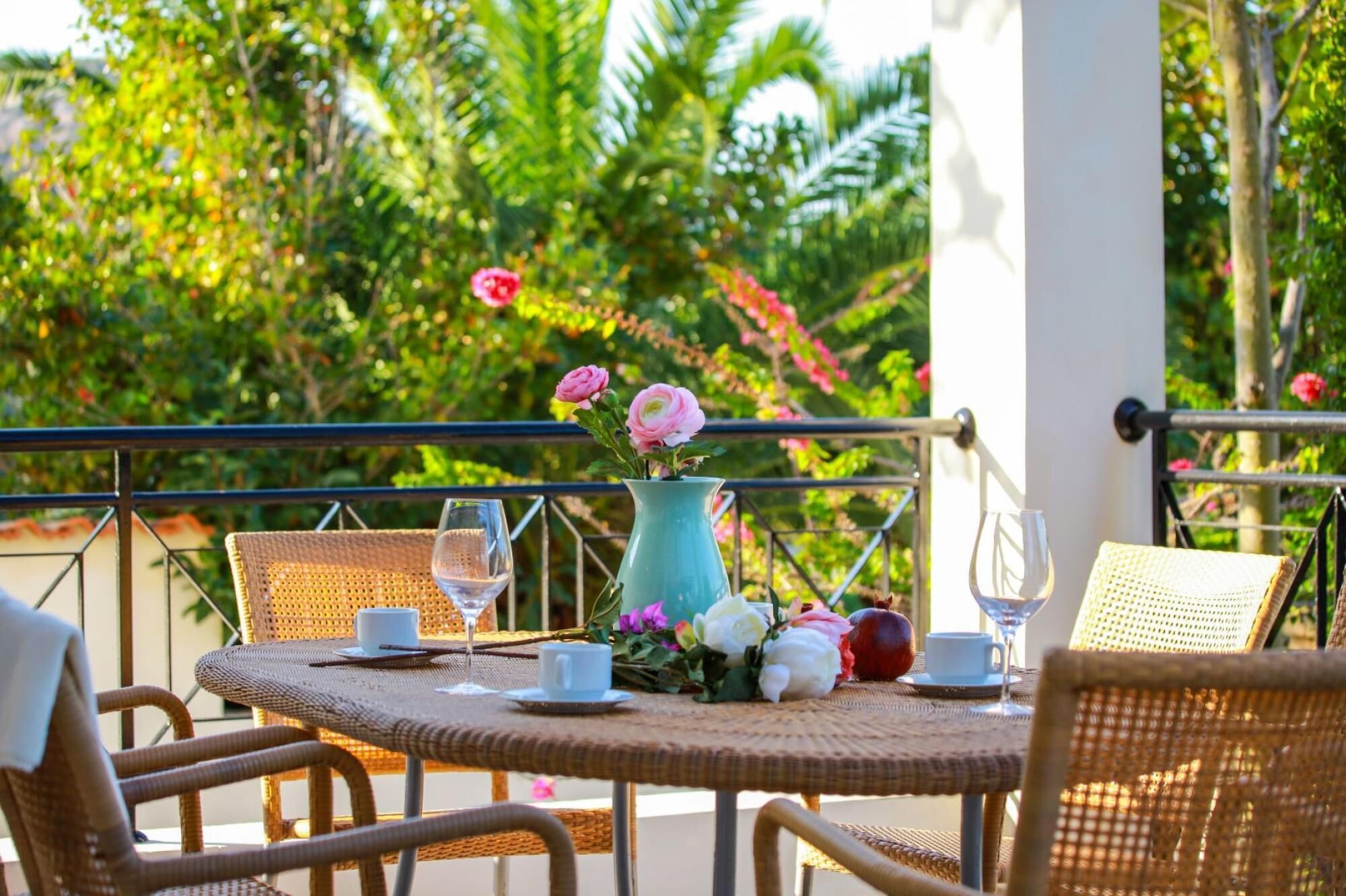 A round table with wicker chairs, a vase with flowers, and two wine glasses on a balcony with a view of lush greenery.