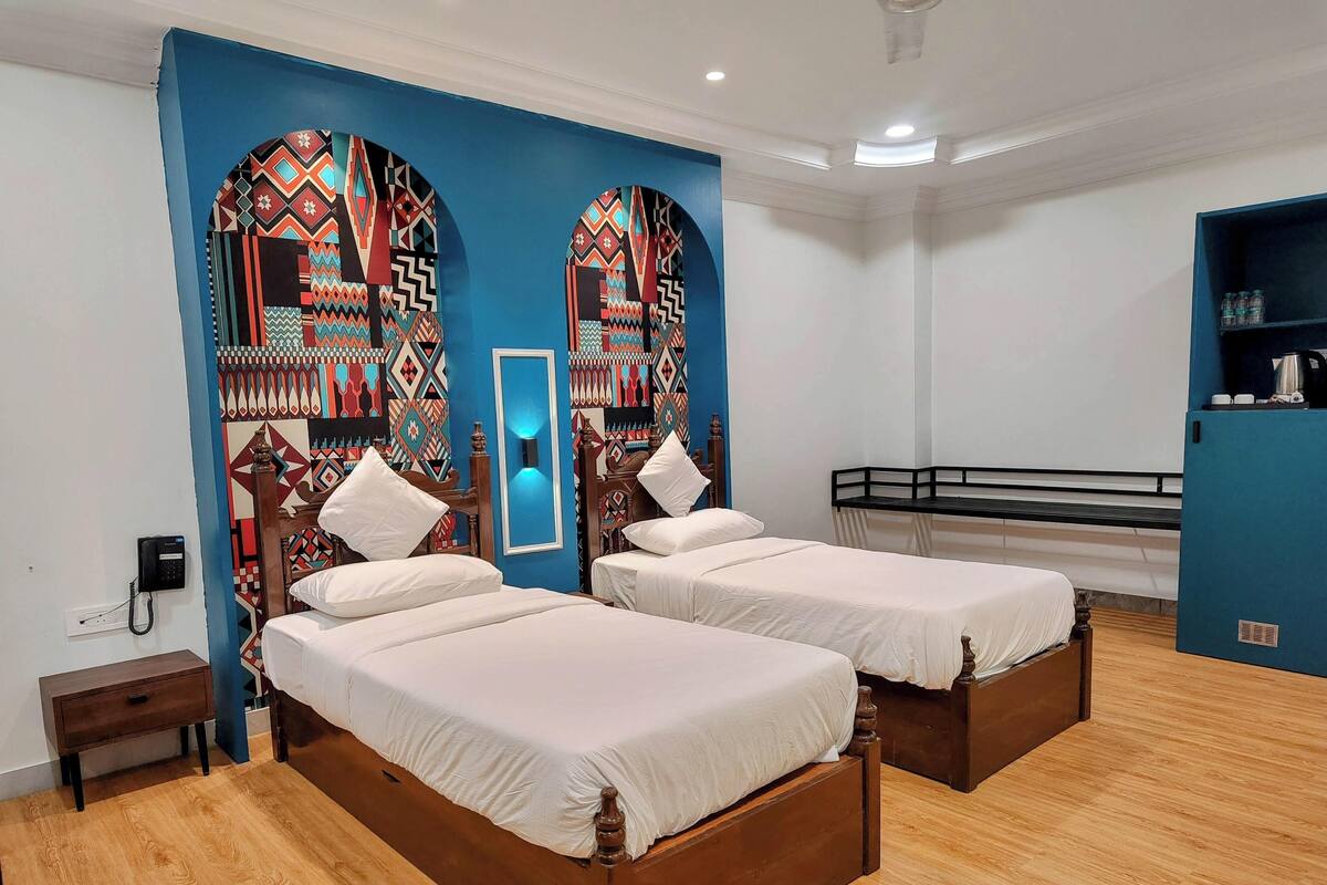 A hotel room with two wooden beds, a blue accent wall, a small bedside table, a nightstand, and a blue cabinet with bottles.