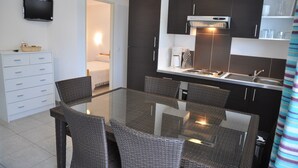 Une cuisine moderne avec des meubles de couleur sombre, une table à manger avec un plateau en verre, des chaises en osier, un téléviseur encastré et une machine à café.