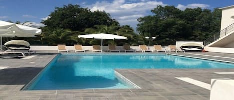 A rectangular swimming pool with a tiled deck, surrounded by lounge chairs and umbrellas.