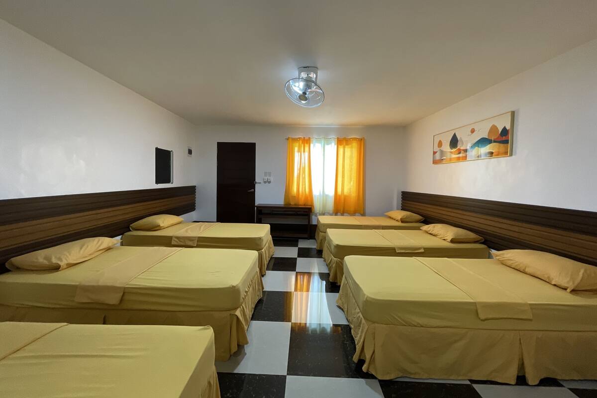 Habitación de hotel con cuatro camas alineadas, piso a cuadros blanco y negro, ventilador de techo y un cuadro en la pared.