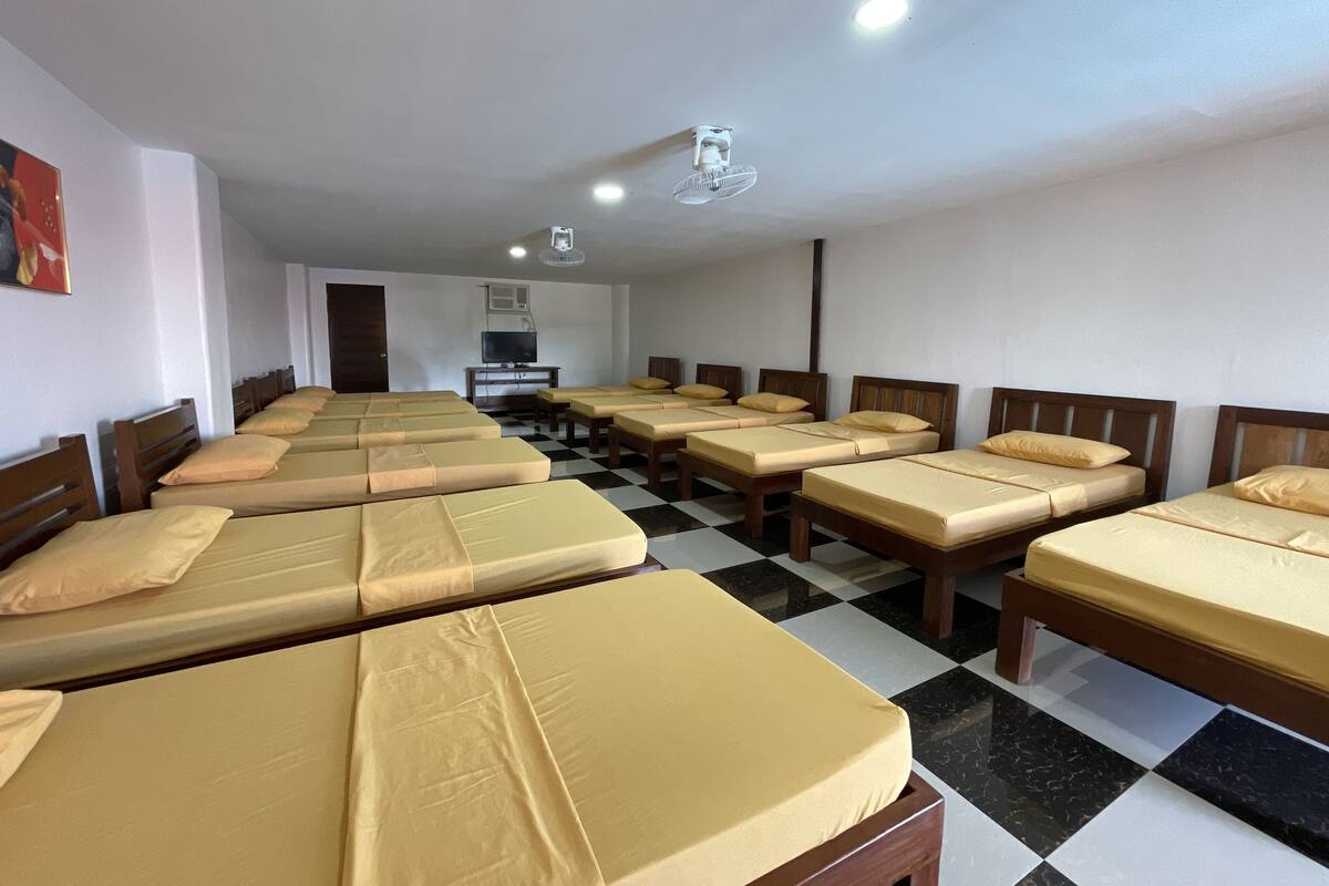 Habitación de hotel con varias camas dispuestas en filas, un televisor sobre un soporte y un ventilador de techo.