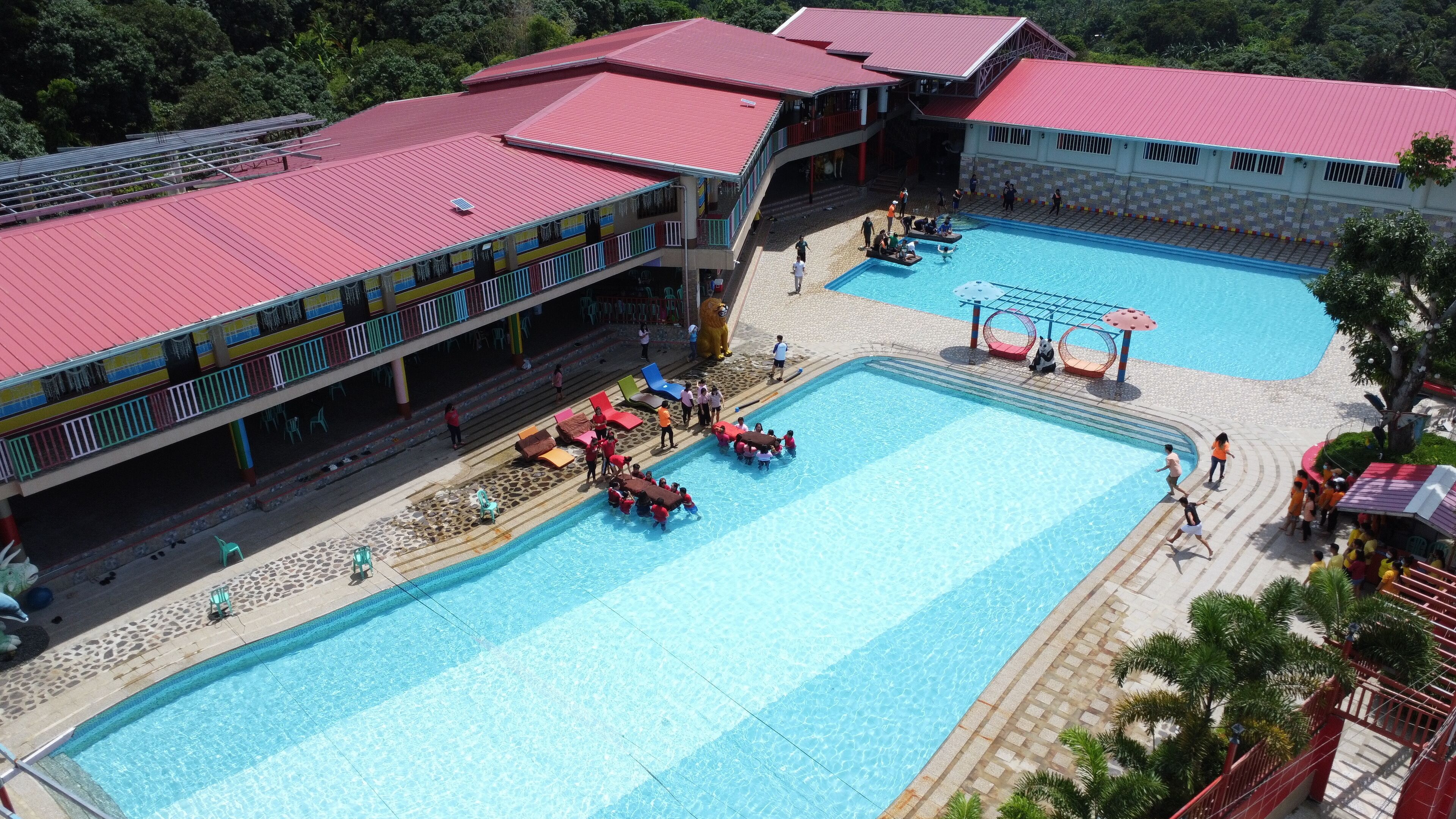 A large outdoor swimming pool with multiple lanes, surrounded by a covered area with red roofs and a seating area with lounge chairs.