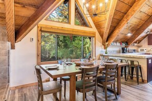 A dining area with a large window, wooden ceiling beams, and a kitchen in the background.