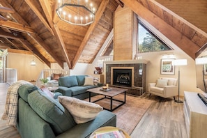 A spacious loft with a high ceiling, a fireplace, and large windows offering a view of the outdoors.