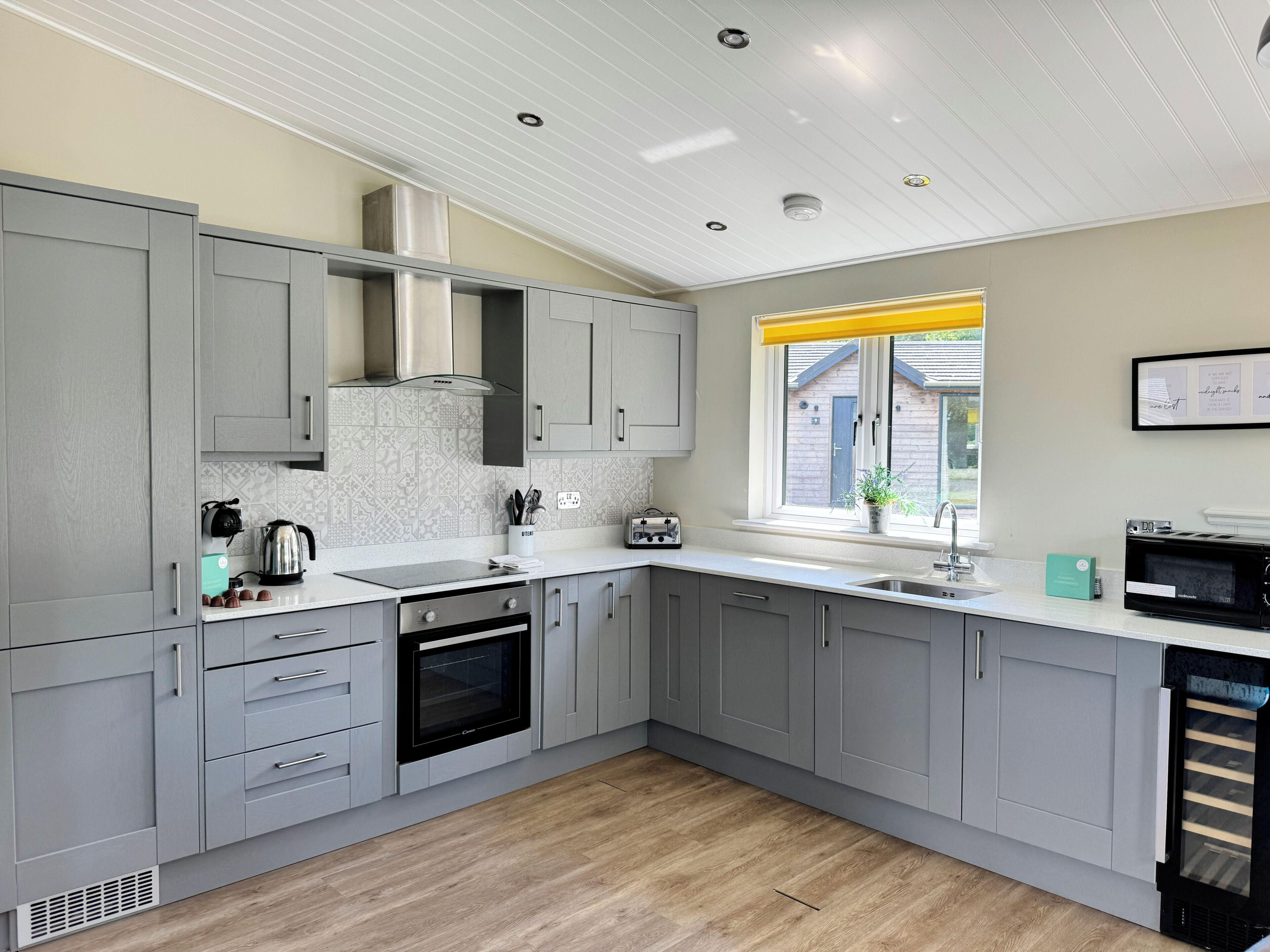 A modern kitchen with grey cabinets, a built-in oven, a microwave, and a window with a yellow blind.