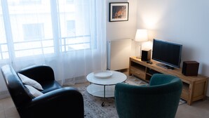 A modern living room with a black leather sofa, a teal armchair, a small round coffee table, a flat-screen TV, and a lamp on a wooden side table. - Cosy, comfortable apartment in the city center (Caen)