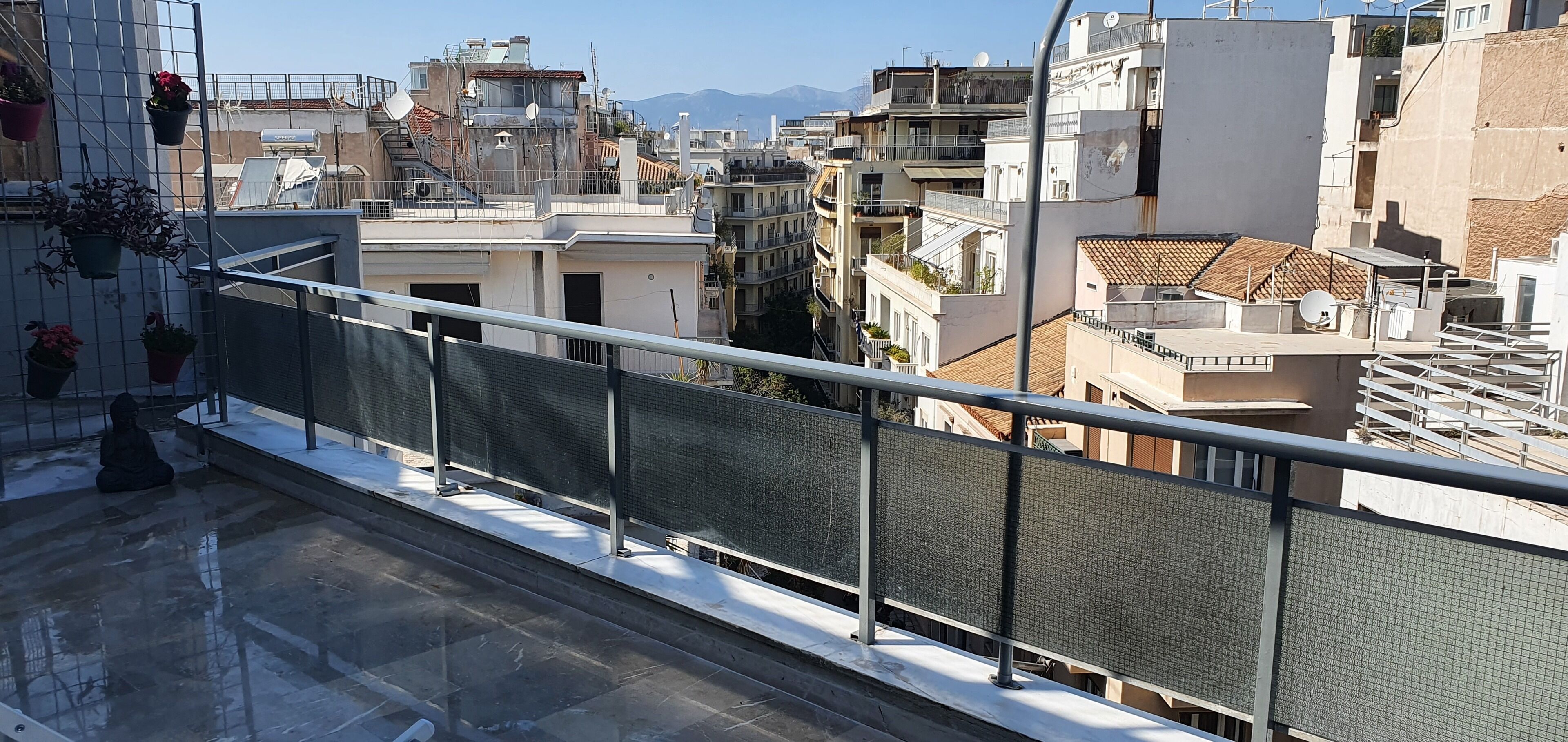 Una terraza en la azotea con barandilla metálica, plantas en macetas y vistas de edificios circundantes.