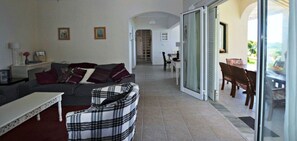 A living room with a sofa, a plaid armchair, a dining area with a table and chairs, and a sliding glass door leading to an outdoor area. - SEASIDE VILLA - HALIKOUNAS with private steps onto the beach (Corfu)