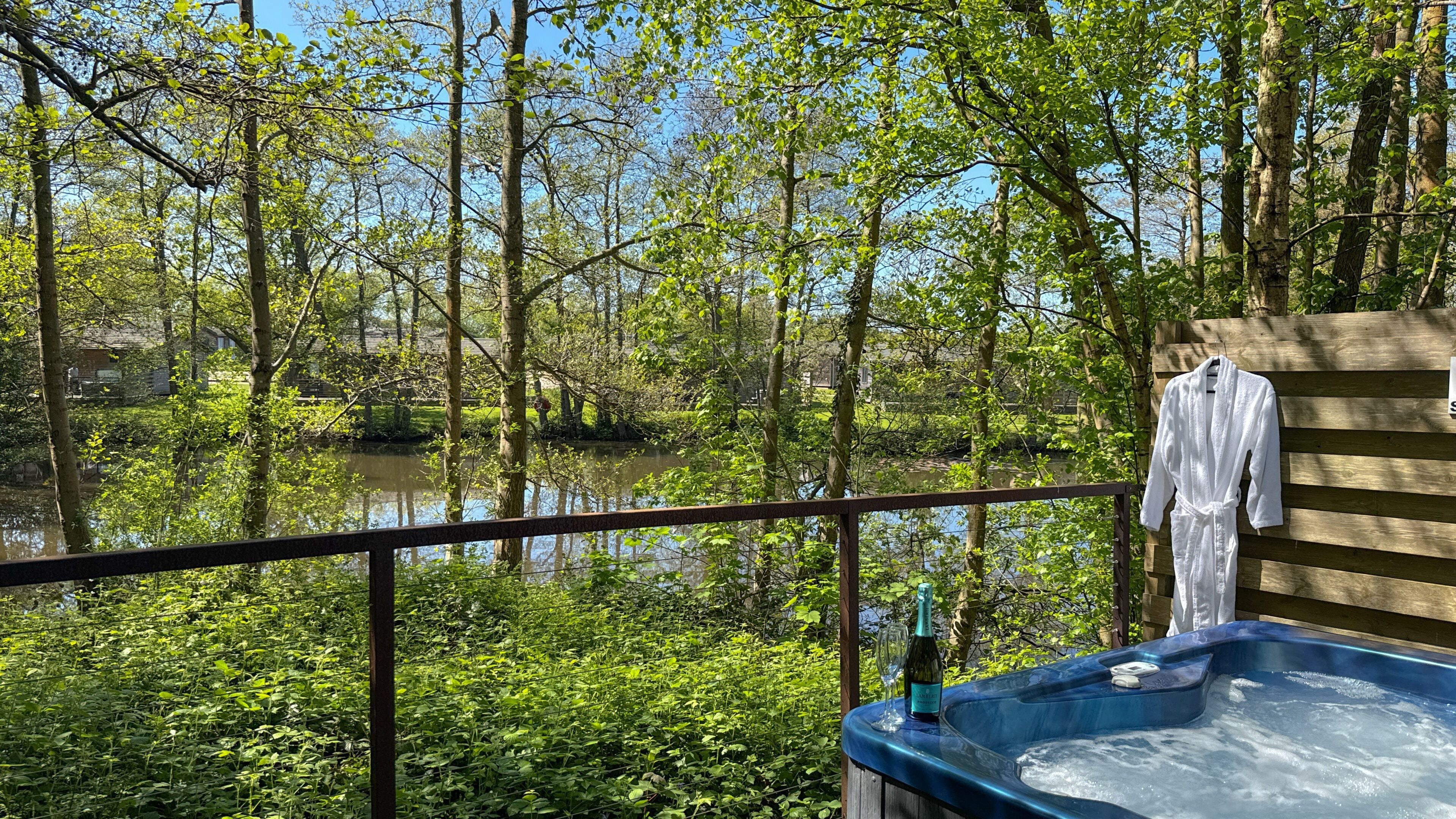 A hot tub with a bottle of wine and a glass, a white robe hanging on a wooden railing, and a view of a forested area with a body of water.