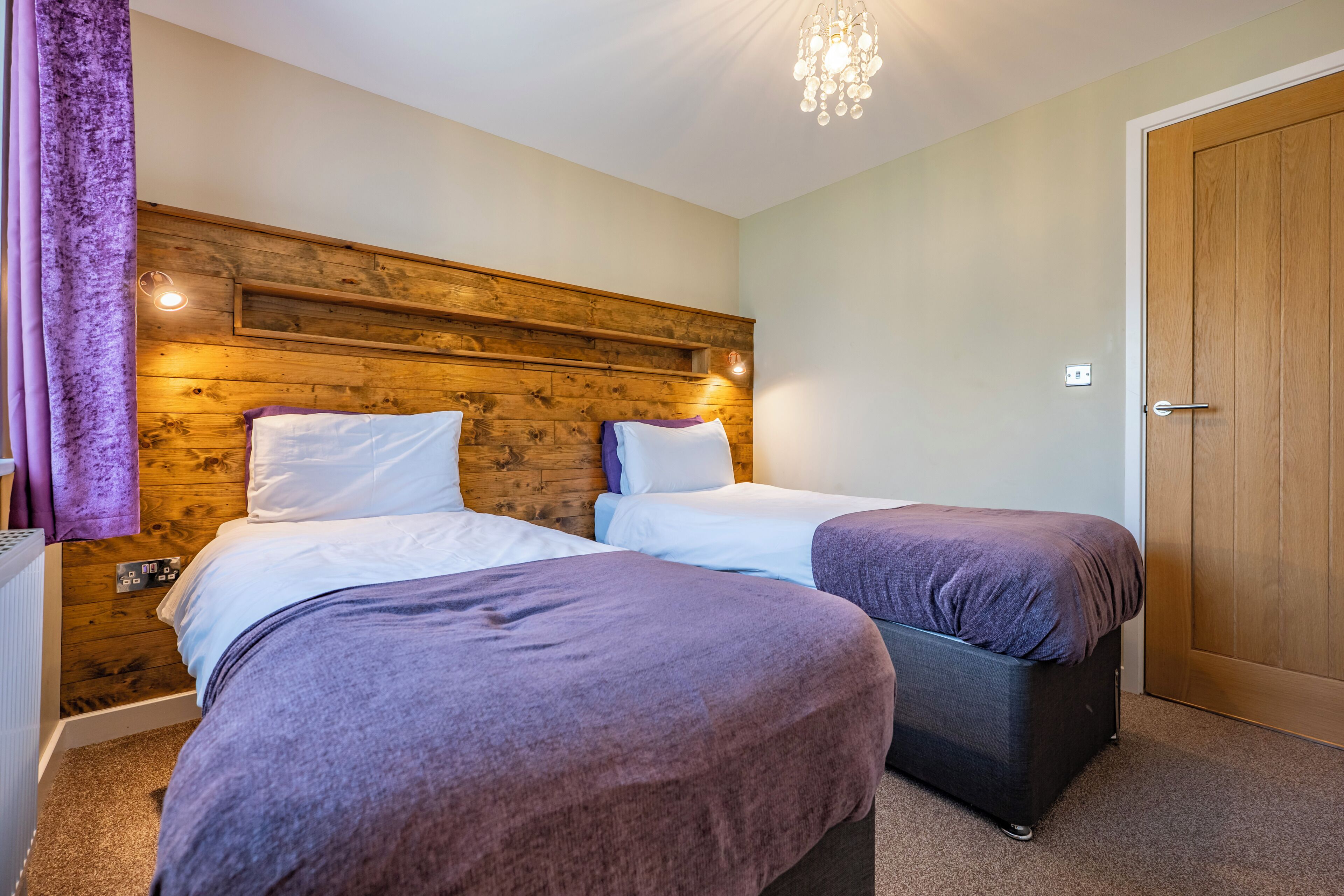 A room with two beds, wooden headboard, a door, and a light fixture.