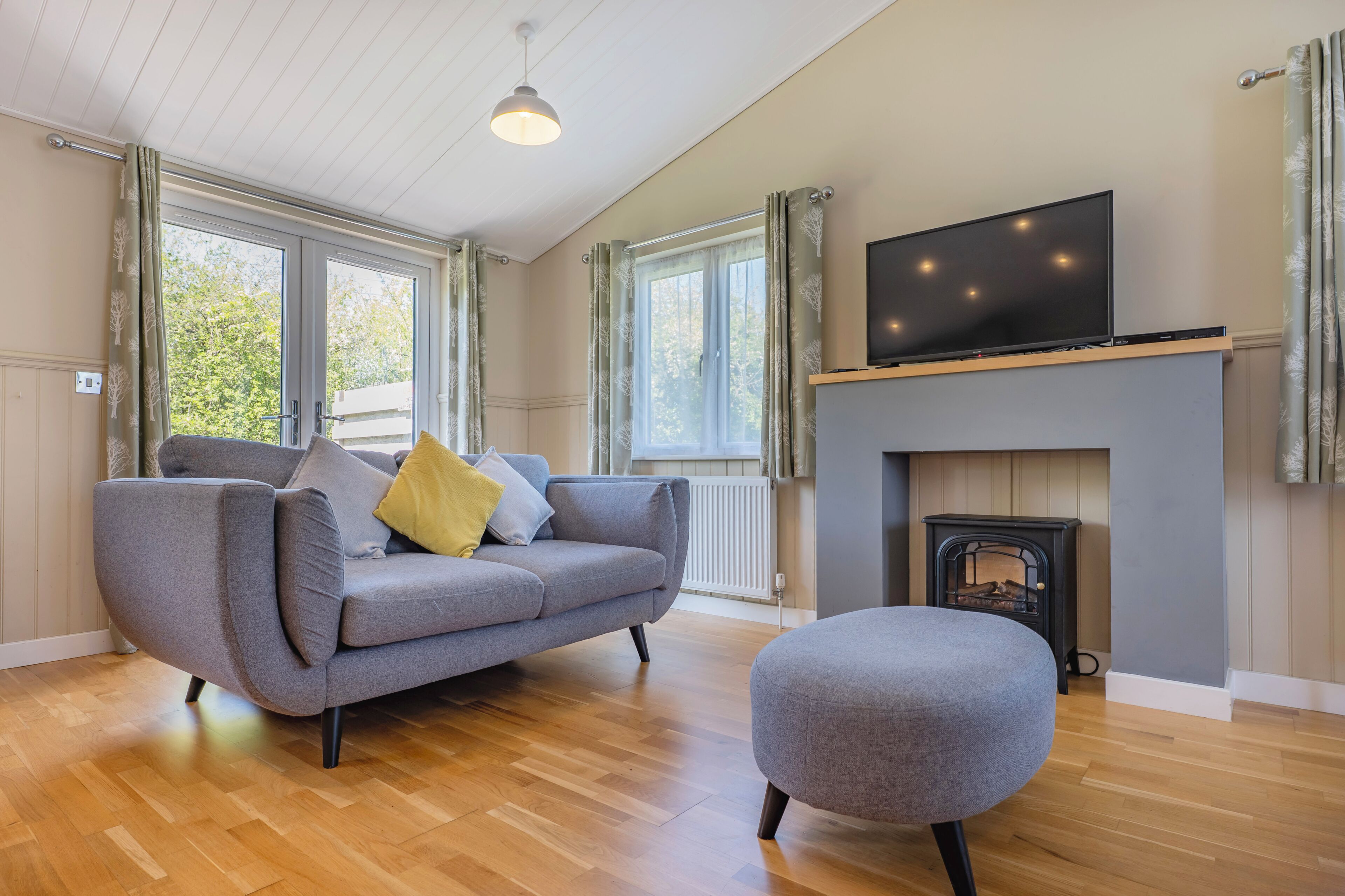 A modern living room with a sofa, ottoman, fireplace, television, and large windows.