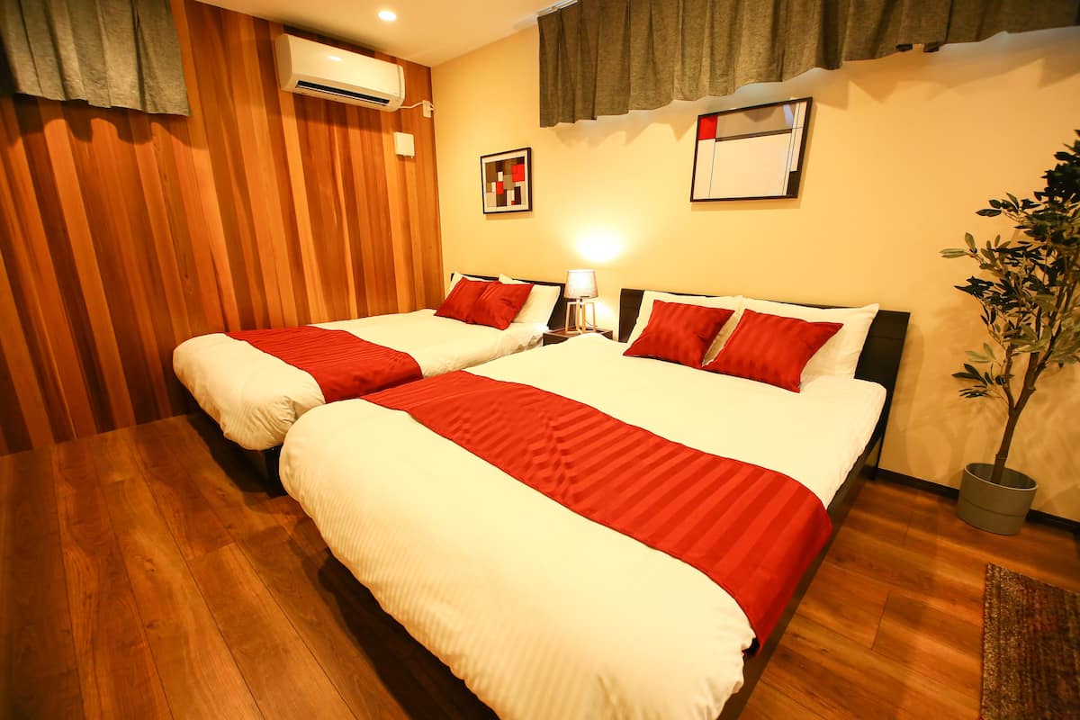 A hotel room with two beds, wooden flooring, an air conditioner, a nightstand with a lamp, and a potted plant.