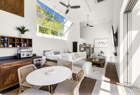 A spacious living area with a white sofa, a dining table with chairs, a kitchen area with a radio, and a large window with a view of trees.