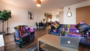 Ein modernes Wohnzimmer mit einer Couch und einem Sessel, bezogen mit farbenfroher Stoff. Es gibt einen Essbereich mit Tisch und Stühlen und eine Küche im Hintergrund.