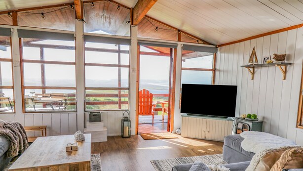 A spacious living area with a large window, a flat-screen TV, and a cozy seating arrangement. - Top Of The World Lodge (Aberdovey)