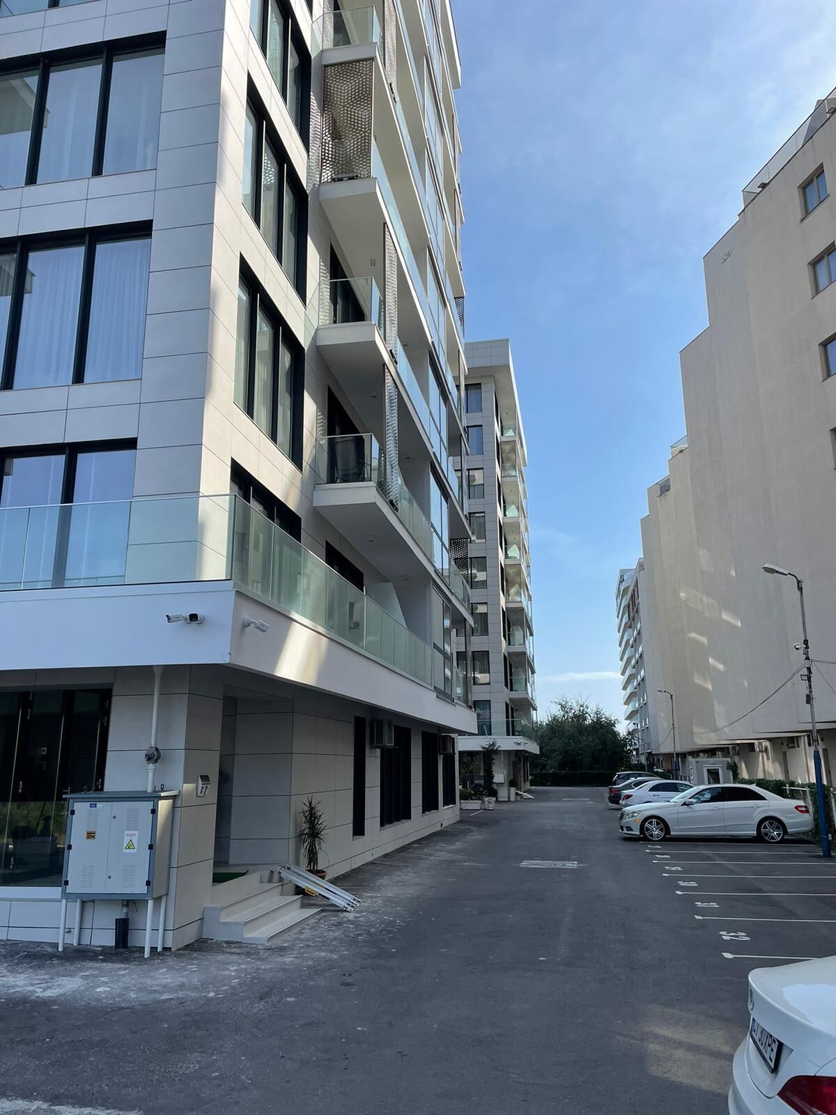 A multi-story residential building with balconies, a parking area, and a clear sky.