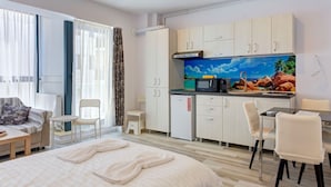 A modern hotel room with a bed, a small desk, a kitchenette, and a large window with curtains.