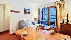 Een eetruimte met een houten tafel, stoelen en een bank. Op de tafel staan kommen en een karaf. Een raam met gordijnen is zichtbaar, en er hangt een foto aan de muur.